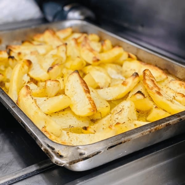 A metal tray filled with golden-brown roasted Greek potatoes, seasoned with herbs and spices.