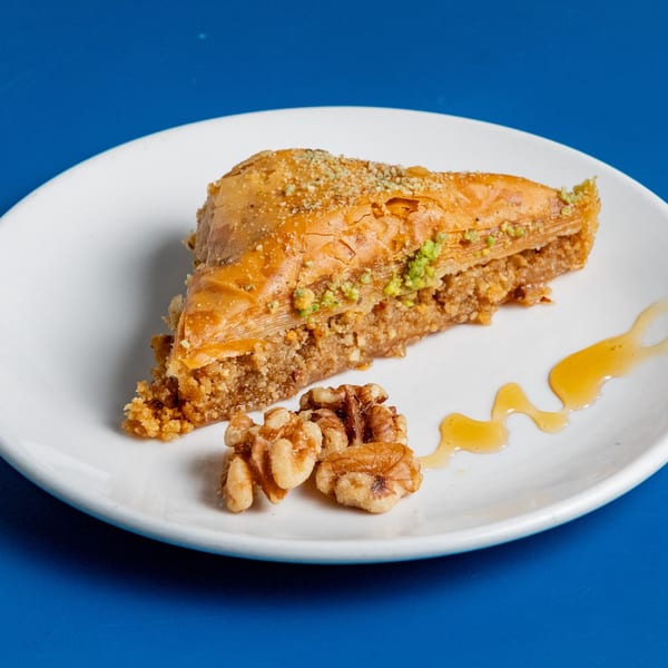 A slice of baklava with walnuts and honey drizzle on a white plate against a blue background.