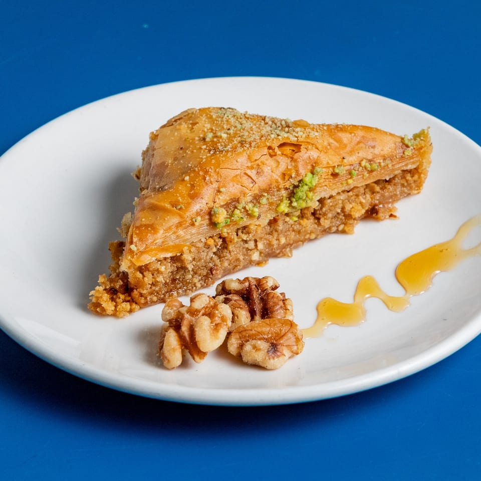 A slice of baklava with walnuts and honey drizzle on a white plate against a blue background.