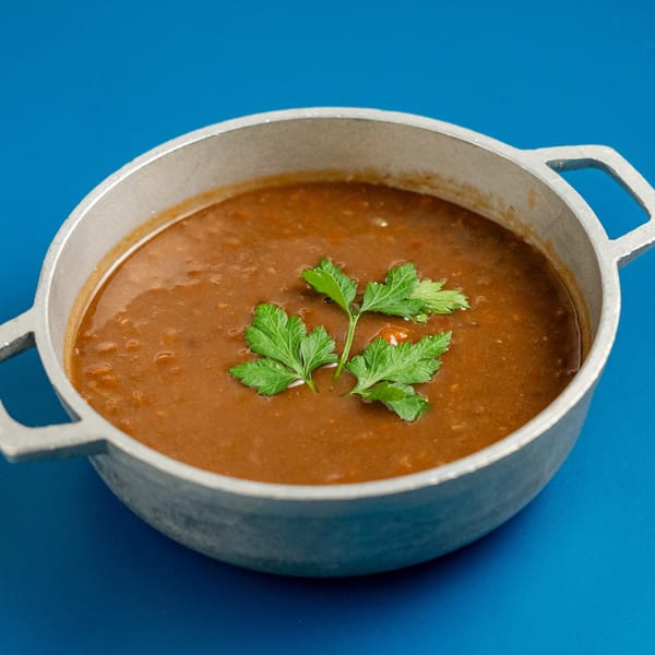 Hearty Greek lentil soup, Fakes, in a metal pot, garnished with fresh parsley on a blue background.