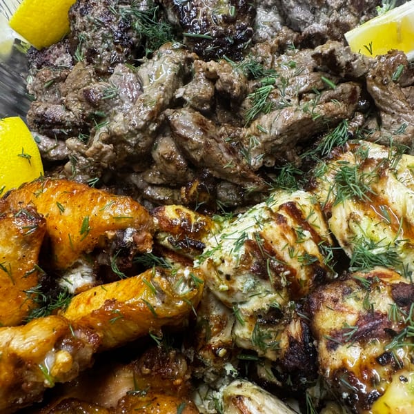 Close-up of grilled chicken wings, chicken pieces, and beef, garnished with fresh dill and lemon wedges.