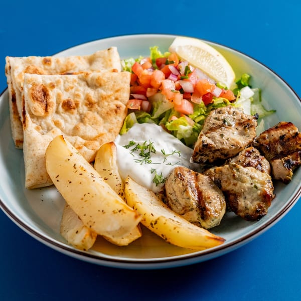 Pita Press Greek chicken souvlaki plate with pita, fries, tzatziki, and salad on a blue background.