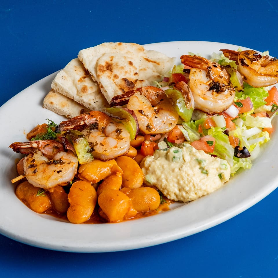 Grilled shrimp skewers, giant beans, hummus, salad, and pita bread on a white plate.
