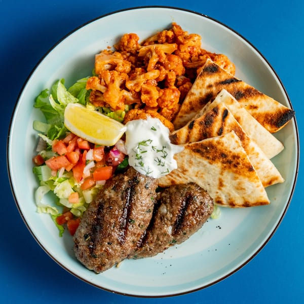 A Greek platter with grilled kofta, pita bread, roasted cauliflower, salad, and tzatziki sauce.