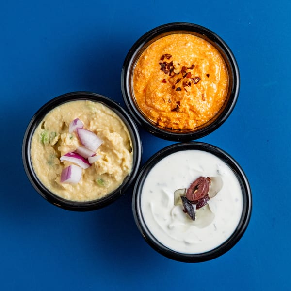 Three Greek dips: hummus with red onion, spicy feta, and tzatziki with olives, on a blue background.